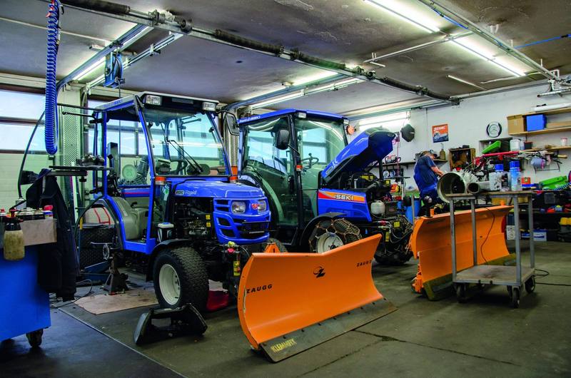 Zwei blaue Iseki-Kompakttraktoren mit Schneepflügen in der Werkstatt, einer mit offener Motorhaube