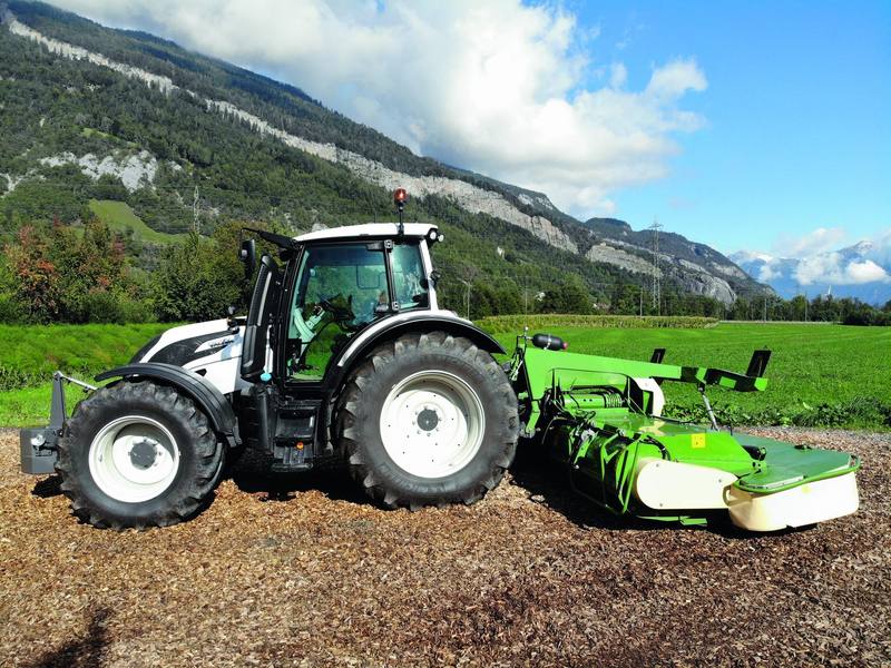 Weisser Valtra-Traktor mit grünem Scheibenmähwerk, im Hintergrund der Bündner Hang