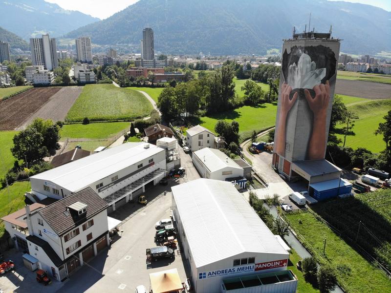 Luftaufnahme der Rheinmühle in Chur: Hallen und Wohnhaus, daneben der Silo-Turm mit dem grossen Wandbild zweier Hände, im Hintergrund die Stadt Chur